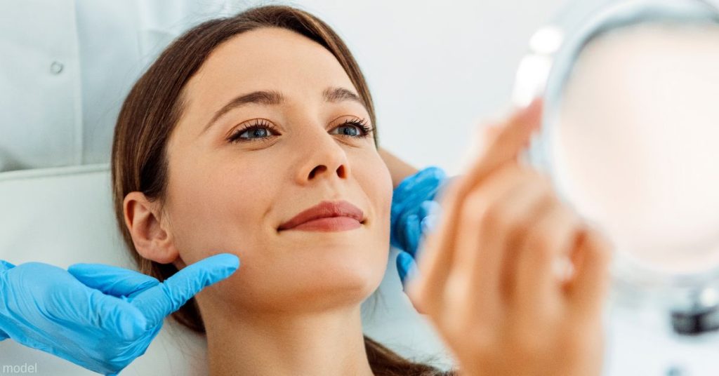 Woman looking in mirror with gloved hands touching her face. (MODEL)