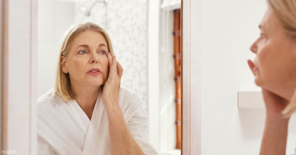 Mature woman looking in the mirror and touching her face. (MODEL)